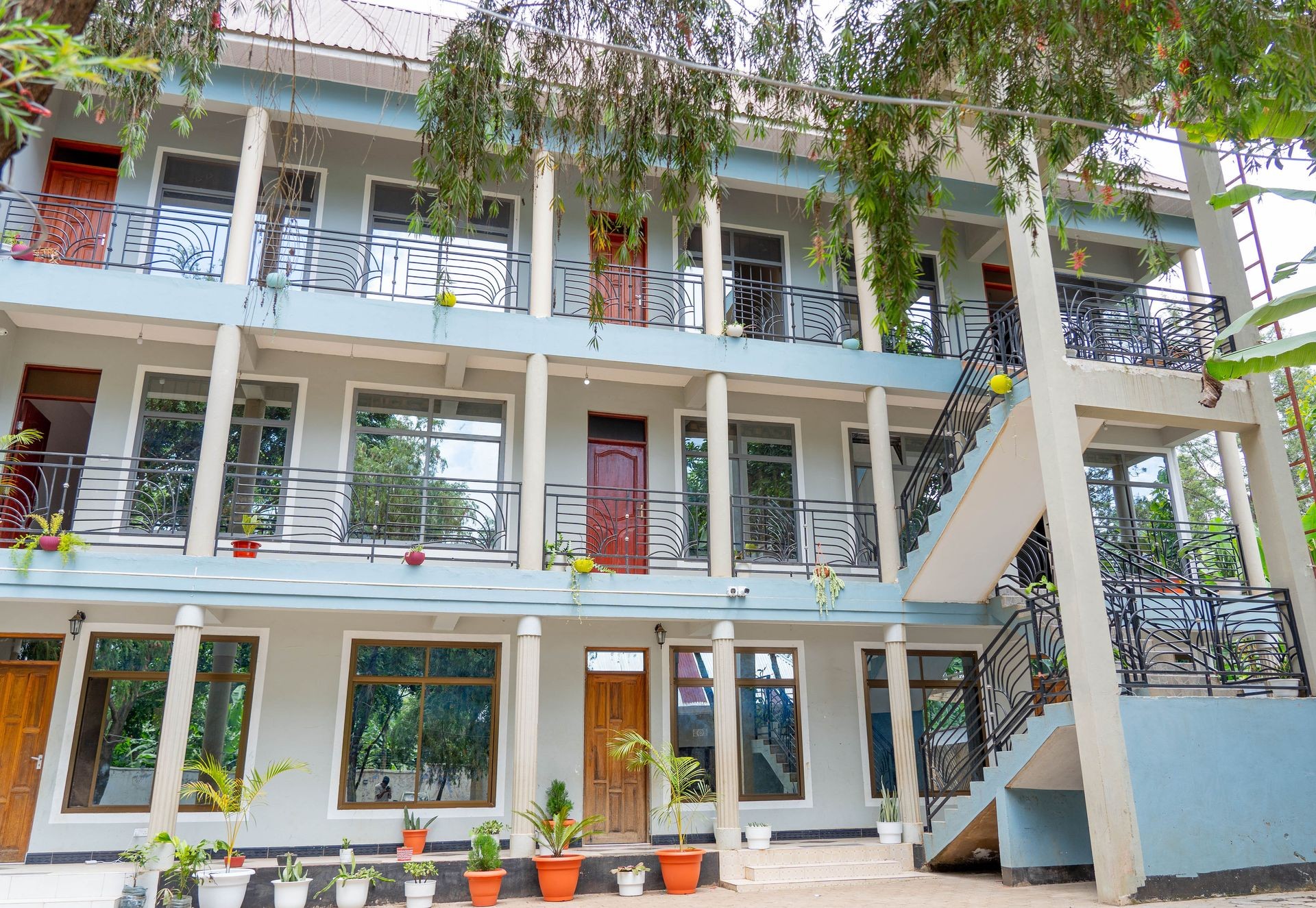Three-story building with multiple doors and balconies, surrounded by potted plants and greenery.
